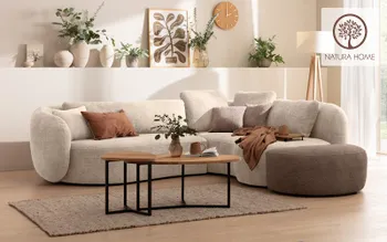 Wohnzimmer im Naturlook: Eine große, helle Rundcouch mit vielen Kissen dominiert den Raum. Naturmaterialien, gedeckte Farben, Holztisch und Vasenarrangement schaffen eine behagliche Wohlfühlatmosphäre. Rechts oben das Natura Home Logo.